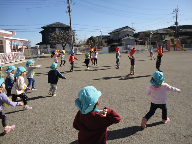 園庭にいる鬼のお面を付けた年長児に豆をまく3歳児の写真。
