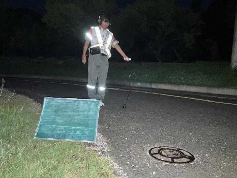 道路に埋設されている水道管を夜間調査している写真です。調査員はヘッドホンをつけ、路面に機器をあてながら歩いて調査しています。