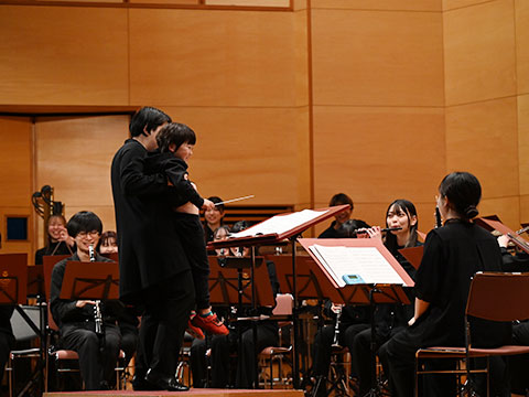 指揮者体験するため抱っこされている男の子を見て笑顔で演奏するオーケストラ