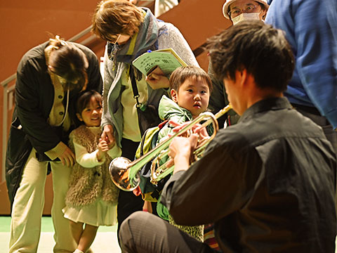 楽器体験でお兄さんが吹くトランペットに興味津々の男の子、恥ずかしがって親の背に隠れてはにかみながら見る女の子