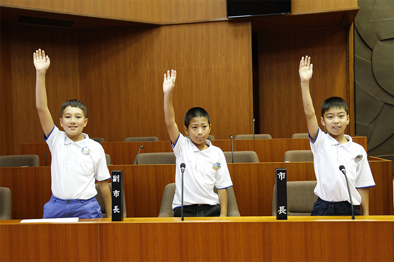 議席に座って元気に手をあげている3人の少年