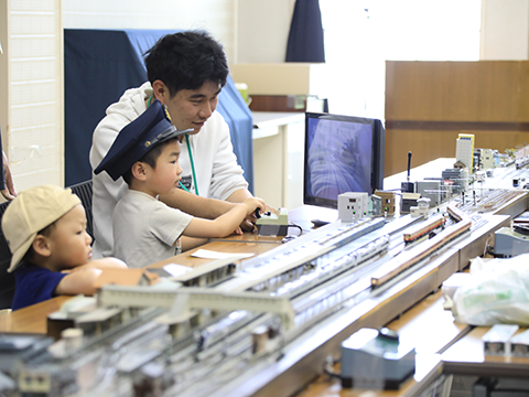 車掌帽をかぶり、大人に教えてもらい鉄道模型を操作する子ども