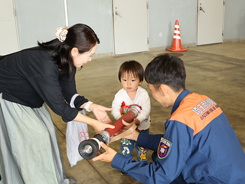 お母さんと消防士さんに支えられ消化用ホースを持つ子ども